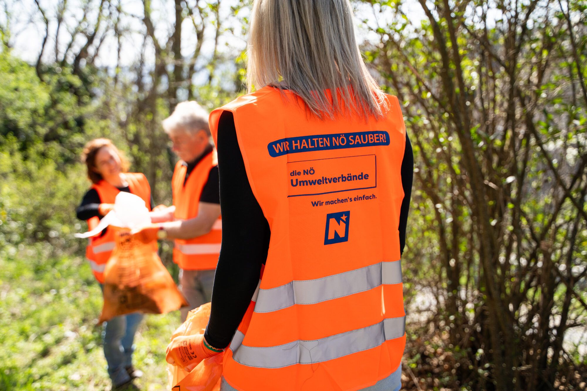 Drei Personen, die mit Fr&uuml;hjahrsputz-Warnwesten bekleidet sind, sammeln in der Natur Abf&auml;lle auf.