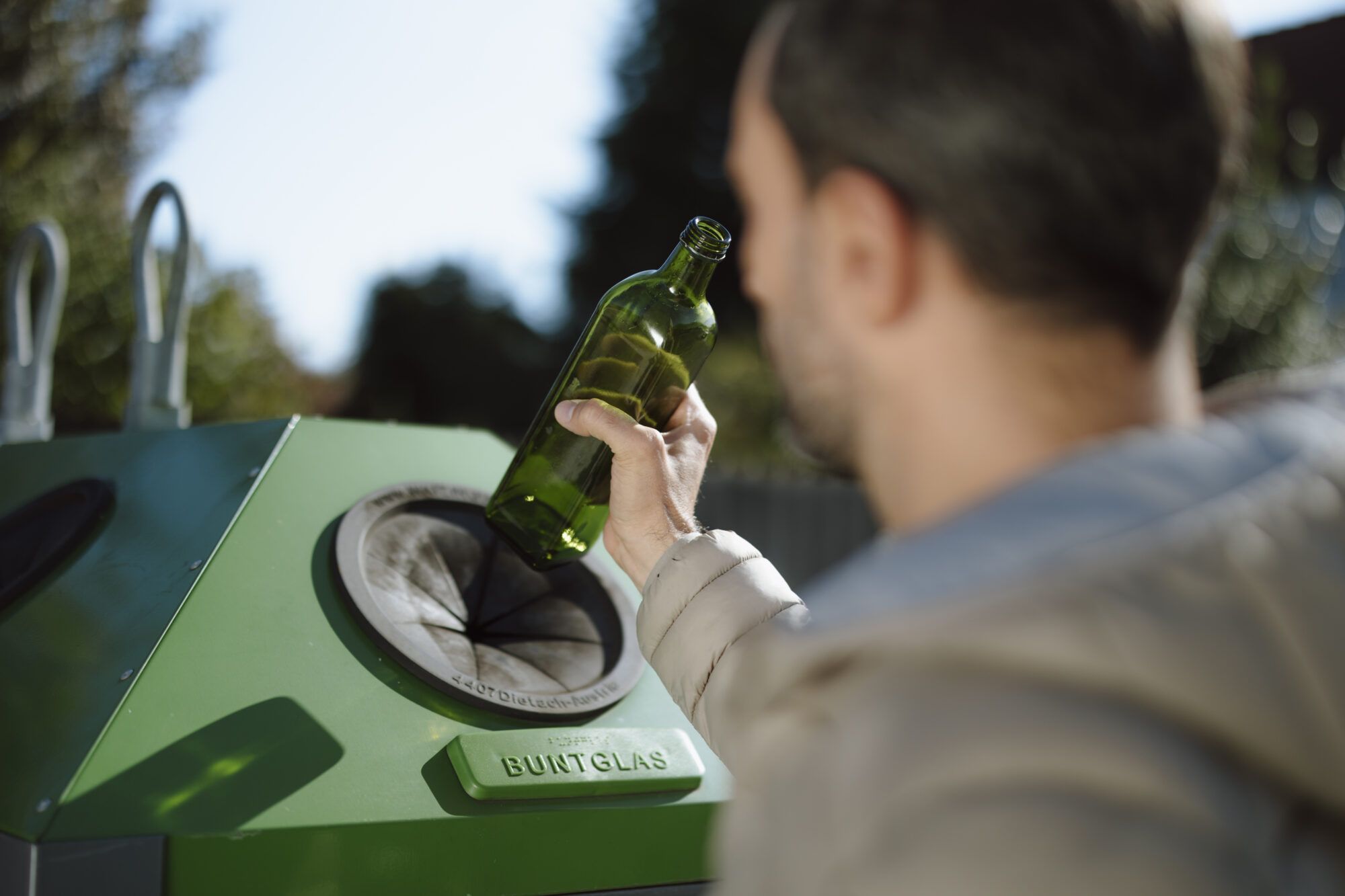 Ein Mann mit braunen Haaren und kurzen Bart ist verschwommen im Vordergrund zu erkennen. Er hält eine grüne Glasflasche in der Hand. Unter der Flasche ist eine schwarze runde Öffnung von einem grünen Buntglas Container zu erkennen.