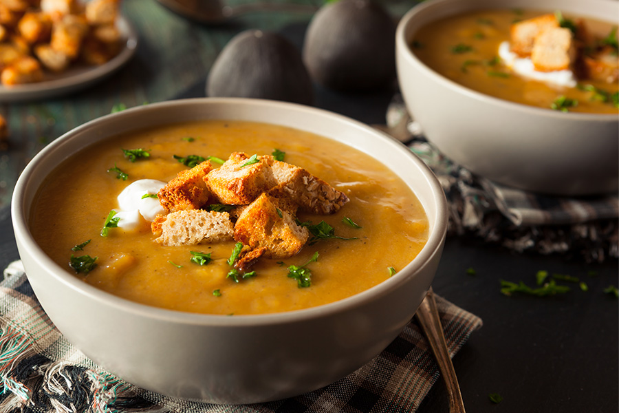 Cremige Kürbissuppe mit Croutons und Kräutern in weißer Schüssel auf grau-weiß karierten Tuch auf dunklem Grund. Im Hintergrund ist unscharf noch eine Schüssel zu sehen.