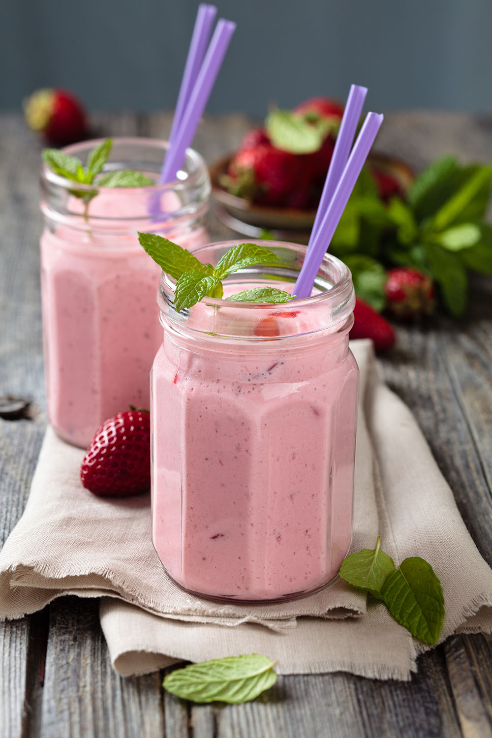 Zwei offene Schraubgläser gefüllt mir hellrosa Erdbeerdrink, garniert mit Minzblättern und Erdbeestücken. Je zwei lila Strohhalm ragen aus den Gläsern. Im Hintergrund unscharf Erdbeeren