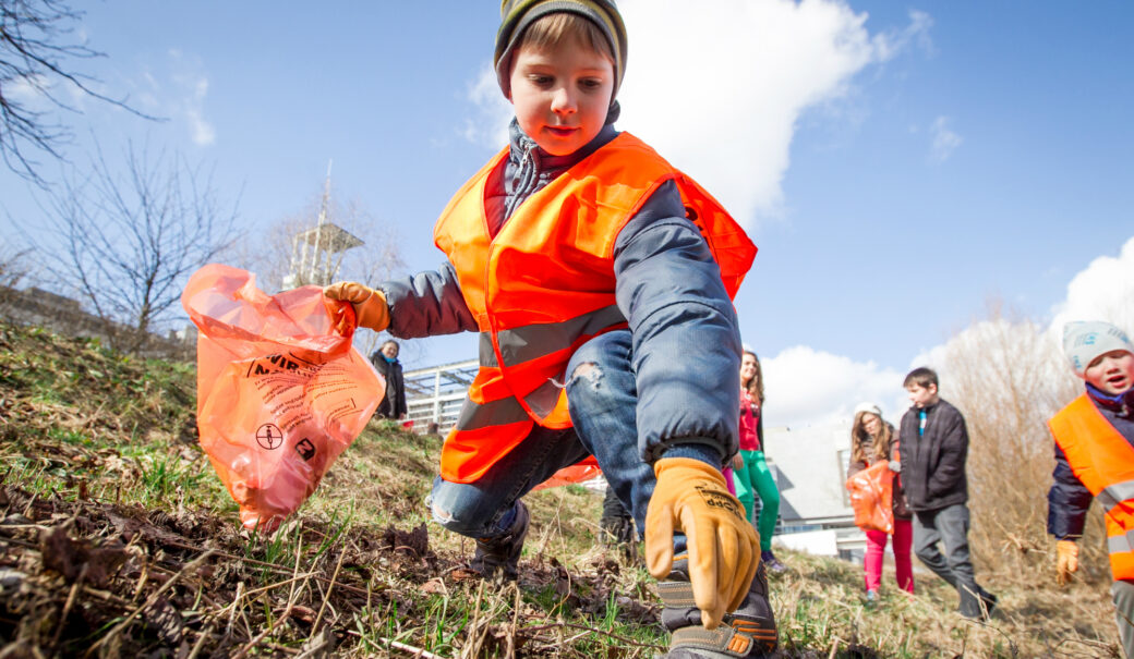 Zu sehen sind Kinder, die mit Warnwesten, Handschuhen und Sammelsäcken ausgestattet sind, Abfälle aus der Natur aufsammeln.